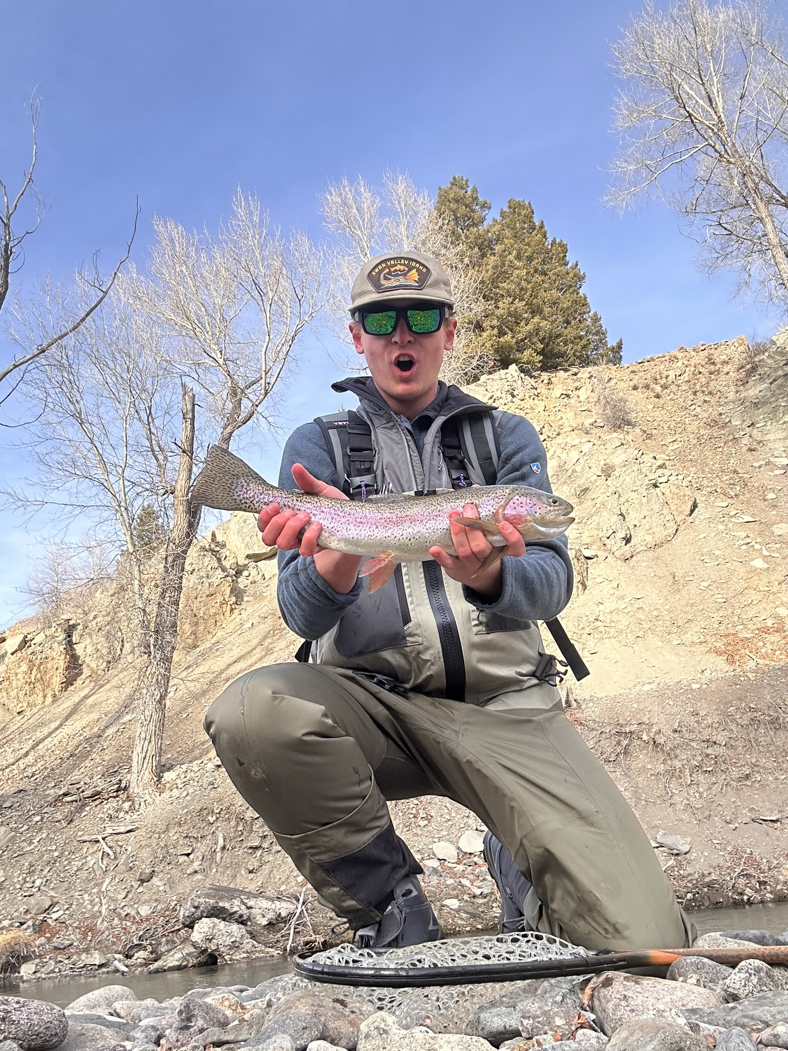 A fly fisherman kneeling on a riverbank in daylight, holding a rainbow trout