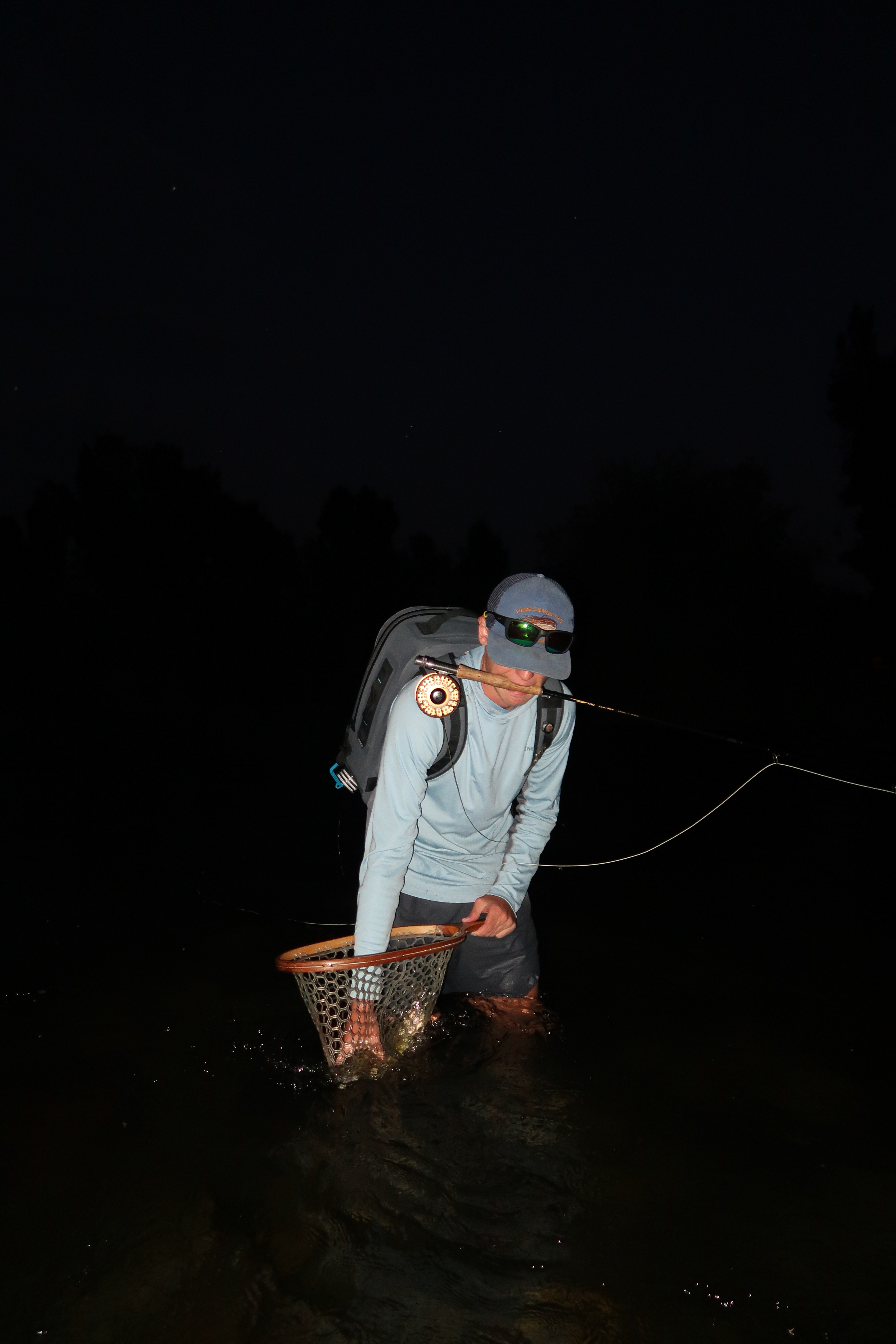 A fly fisherman netting a catch at night, headlamp cutting through the dark water